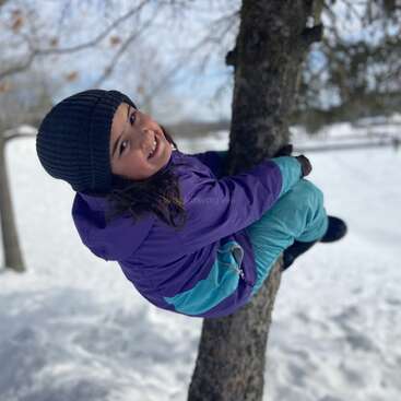Une jeune fille, vêtue d'une veste violette, d'un pantalon de neige sarcelle et d'un bonnet noir, sourit en grimpant à un arbre dans un cadre extérieur enneigé.