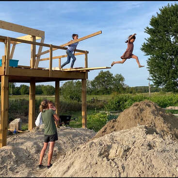 The image depicts a group of people working on a wooden structure, with one person jumping off the side and another carrying a long piece of wood.