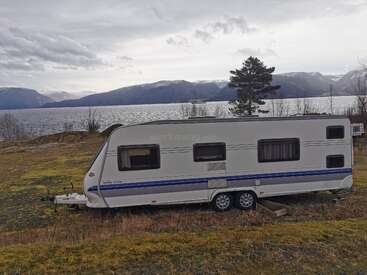 A white caravan is parked on grassy land overlooking a scenic lake, surrounded by mountains and trees under a cloudy sky, creating a peaceful nature retreat.
