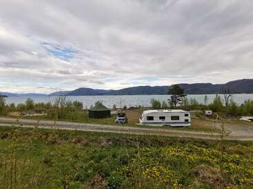A scenic lakeside campsite with a caravan, tent, and car. Mountains and cloudy skies reflect on the calm water, surrounded by greenery and wildflowers.