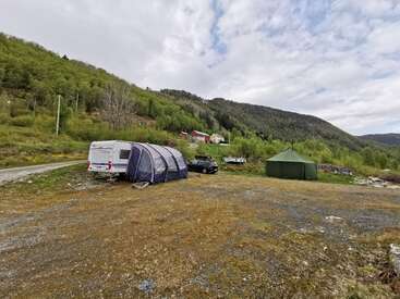A campsite with a caravan, tent, and car is set on open ground surrounded by green hills, trees, and distant houses under a cloudy sky.