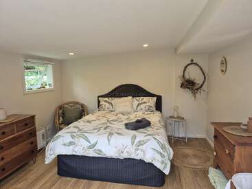 Este quarto aconchegante tem uma cama de casal com roupa de cama floral, duas cômodas de madeira, uma janela pequena, uma cadeira de vime, uma guirlanda decorativa, iluminação natural e piso de madeira.