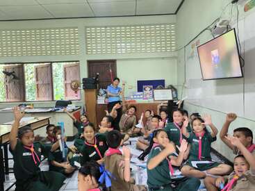 A lively classroom scene with cheerful students sitting on the floor, waving and smiling at the camera, while a teacher stands near the back, overseeing activities.