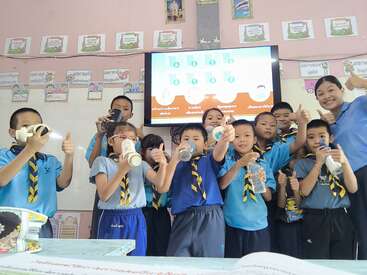 A group of young students and their teacher in uniforms smile and hold water bottles, giving thumbs up, standing in a classroom with a digital screen behind them.
