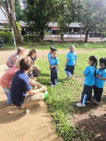 A group of young children in blue uniforms interacts with three adults outdoors, surrounded by greenery, trees, and a school building in the background.