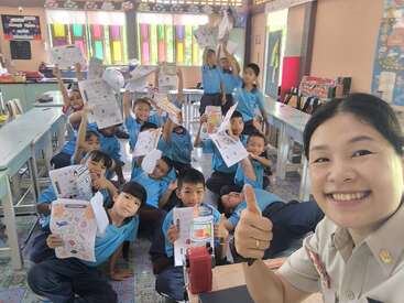 A smiling teacher takes a selfie in a colorful classroom, while happy students in blue uniforms proudly display their worksheets and smile enthusiastically toward the camera.