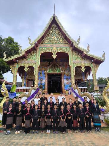 A group of children and adults wearing traditional clothing poses in front of a beautifully ornate, gold-decorated temple with elaborate architectural details and naga sculptures.