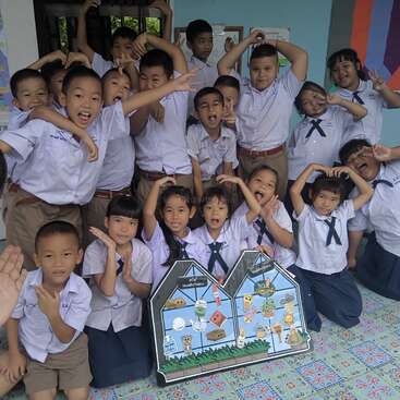 A group of cheerful school children in uniforms pose together indoors, making heart shapes and playful gestures, with colorful educational boards displayed in front of them.