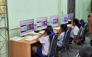 In a classroom, several young students in uniform sit at desks using desktop computers. They appear focused, engaging with educational content displayed on the screens.
