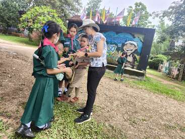 A group of young children in scout uniforms gather around an adult outdoors. The adult gives instructions while another child stands alone near a colorful sign.