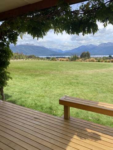 Eine ruhige Holzveranda bietet einen Blick auf einen üppigen grünen Rasen mit Weinreben über dem Kopf, der zu einem ruhigen See und majestätischen Bergen unter teilweise bewölktem Himmel führt.