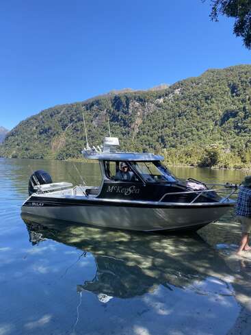 Ein kleines schwarz-silbernes Boot mit dem Namen "McKenSea" schwimmt auf einem klaren, ruhigen See, umgeben von üppig grünen Bergen unter einem strahlend blauen Himmel.