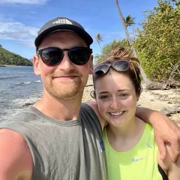 A smiling couple poses for a selfie on a sunny tropical beach. They wear sunglasses and casual clothes, surrounded by palm trees, blue sky, and ocean.