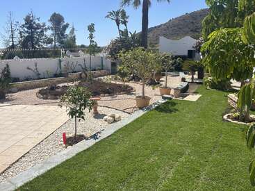 Un jardin bien entretenu, avec une pelouse verte, des plantes en pot, des allées pavées, une terrasse carrelée, des arbres, une clôture blanche et des collines en arrière-plan, sous un ciel bleu limpide.