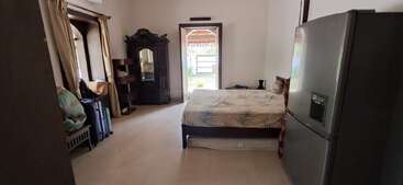 Cette chambre dispose d'un lit, d'un grand réfrigérateur, d'une armoire en bois avec miroir, de fenêtres avec rideaux, de bagages et d'une porte voûtée menant à l'extérieur.