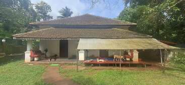 Une maison traditionnelle au toit de tuiles entourée de verdure. Un chien se tient sous le porche, couvert par un hangar. L'atmosphère est tranquille et rurale.