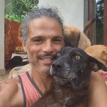 Un homme souriant aux cheveux grisonnants prend dans ses bras un chien noir. Plusieurs autres chiens se trouvent à l'arrière-plan. Ils apparaissent à l'extérieur, profitant d'un moment paisible et heureux ensemble.