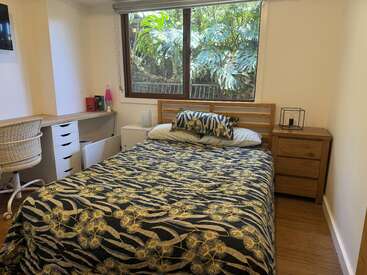 A cozy bedroom featuring a double bed with a leafy-patterned cover, wooden furniture, a study desk with chair, large window, and lush greenery outside.