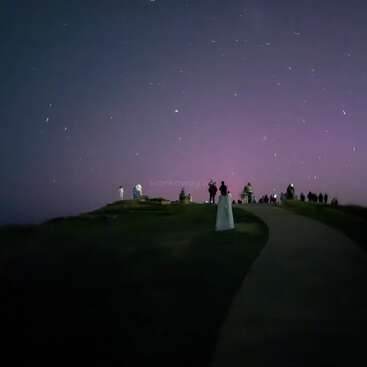 A group of people stands on a hill under a starry night sky, illuminated by a soft purple glow, creating a serene, dreamy atmosphere.