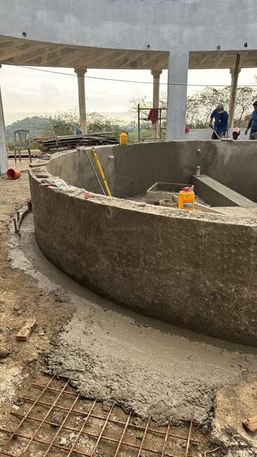 Uma estrutura circular de concreto em construção, possivelmente uma piscina ou tanque. Você pode ver ferramentas, materiais de construção e trabalhadores. O local é ao ar livre com uma vista panorâmica.