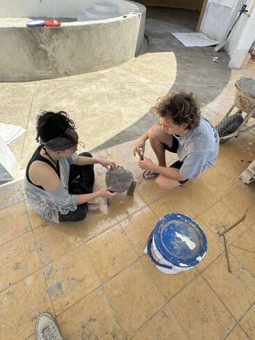 Duas pessoas estão sentadas em um piso de ladrilhos envolvidas em uma atividade prática, possivelmente trabalhando com cimento ou gesso, cercadas por materiais e ferramentas de construção.
