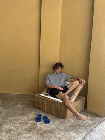 Um homem relaxa em uma cadeira de madeira contra uma parede amarelo mostarda, vestindo uma camiseta cinza e bermuda preta. Chinelos azuis são colocados no piso texturizado.