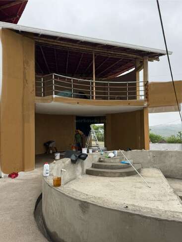 Esta imagem mostra um edifício parcialmente construído ou reformado com uma estrutura externa curva, uma escada e várias pessoas trabalhando sob uma varanda coberta em um dia nublado.