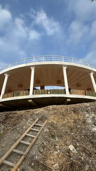 Un pavillon circulaire avec des colonnes blanches et des lumières suspendues se dresse sur une pente. Une échelle en bois s'appuie sur la colline de terre, menant à la structure ouverte.