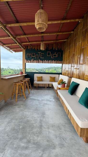 Ce patio confortable en bambou est équipé de bancs rembourrés, de tabourets en bois, de lampes suspendues et d'un panneau "Monte Olón". Il donne sur des collines verdoyantes par une fenêtre ouverte.