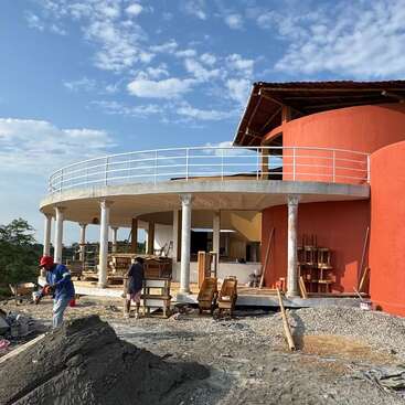 A modern circular building under construction with white pillars and an orange curved wall. Two workers are on site, surrounded by building materials and tools outdoors.