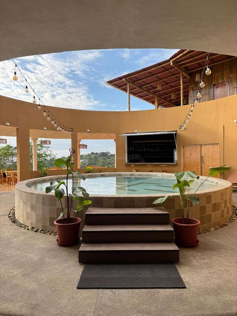 Une piscine circulaire accueillante avec des marches en bois, entourée de plantes en pot. Des guirlandes lumineuses sont suspendues au-dessus, avec un bar et un coin salon adossés à des murs chauds de couleur beige.
