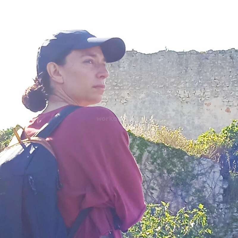 Une femme portant une casquette sombre et une chemise marron se tient debout à l'extérieur avec un sac à dos, regardant pensivement au loin. Derrière elle se trouvent un mur de pierre et de la verdure.