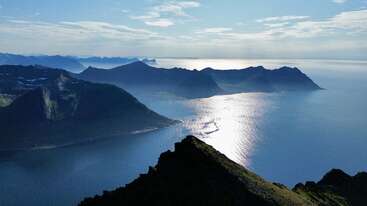 Uma paisagem costeira deslumbrante com picos de montanhas dramáticas, mar azul calmo e a luz do sol refletindo lindamente na água sob um céu parcialmente nublado, criando uma atmosfera serena.