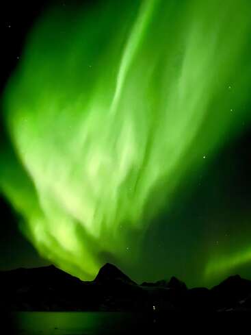 Uma exibição dramática de luzes verdes da aurora boreal dança no céu noturno acima de montanhas escuras e silhuetas, refletindo suavemente em uma água calma e tranquila.