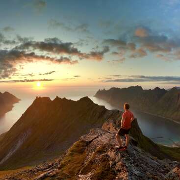 A person stands on a mountain ridge at sunset, overlooking dramatic peaks, a winding coastline, and calm waters, embraced by colorful skies and scattered clouds.