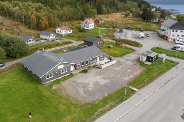 Esta imagem aérea mostra um pequeno vilarejo rural com várias casas, um grande prédio central, área de estacionamento, arredores gramados, árvores e um corpo d'água à distância.