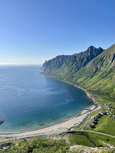 Uma deslumbrante paisagem costeira apresenta uma impressionante cadeia de montanhas, mar azul cristalino, praia de areia, estrada sinuosa, colinas verdes e casas espalhadas sob um céu azul brilhante.