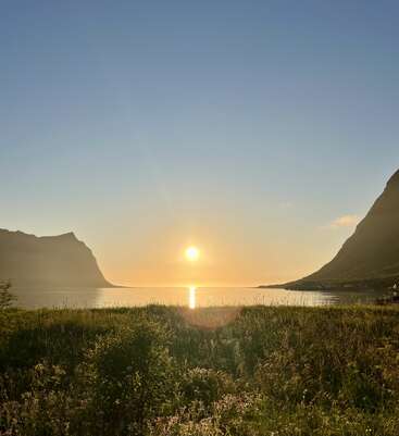 Um belo pôr do sol sobre um lago calmo, ladeado por montanhas e prados gramados em primeiro plano, com um céu azul claro e dourado acima.