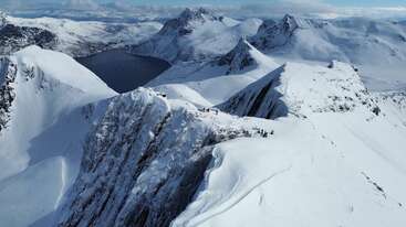Um cume de montanha coberto de neve com vários caminhantes percorrendo a trilha estreita. Cercado por picos impressionantes e um lago azul-escuro aninhado entre as montanhas, sob um céu claro.