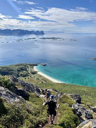 Os caminhantes descem uma trilha rochosa com vista para uma deslumbrante praia azul-turquesa, uma floresta verde e um oceano calmo. Ilhas e montanhas distantes se erguem sob um céu claro e parcialmente nublado.