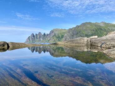 Uma paisagem deslumbrante apresenta montanhas verdes irregulares refletidas perfeitamente em uma piscina de água calma e límpida, com céu azul e nuvens dispersas acima da cena pacífica.