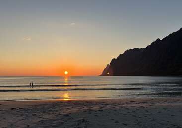 A imagem mostra uma praia tranquila ao pôr do sol, com duas pessoas nadando em águas calmas, montanhas impressionantes à direita e o sol brilhante próximo ao horizonte.