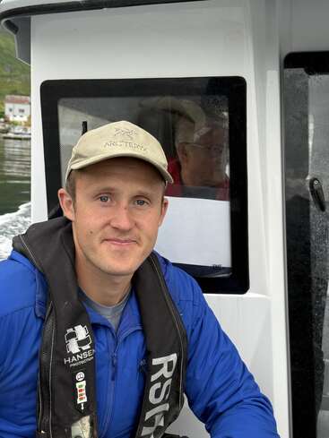Um homem sorridente, com jaqueta azul e boné bege, está sentado em um barco. Ele usa um colete salva-vidas preto. Outra pessoa pode ser vista atrás da janela.