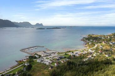 Uma pitoresca vila costeira com casas espalhadas, uma marina e uma vegetação exuberante. O mar azul calmo encontra montanhas distantes sob um céu claro e parcialmente nublado, criando uma beleza serena.