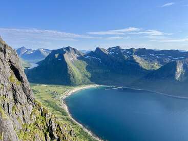 Uma paisagem costeira deslumbrante com água azul clara, montanhas verdes impressionantes, picos cobertos de neve e uma praia de areia sob um céu azul brilhante, visto de cima.