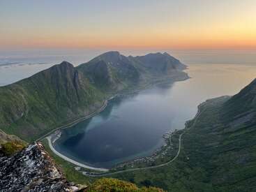 Uma vista aérea impressionante de um fiorde norueguês durante o pôr do sol. Montanhas íngremes e verdes cercam águas azuis calmas, com uma estrada costeira sinuosa e um pequeno vilarejo abaixo.