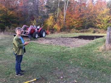 The image depicts a woman standing in a grassy field, with a red tractor and a partially dug garden bed in the background, surrounded by trees with autumn foliage.