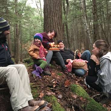 The image depicts a family of five celebrating a birthday in the woods, with a cake and basket on a mossy hill, surrounded by trees and fallen leaves.