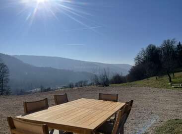 Uma mesa de madeira ao ar livre com seis cadeiras fica no cascalho sob a luz do sol, com vista para colinas enevoadas, montanhas distantes e árvores sob um céu azul claro.