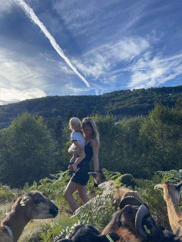 Uma mulher carrega uma criança pequena enquanto caminha entre cabras em uma paisagem verde e exuberante. O céu azul com nuvens dramáticas e rastros se estende acima de você. Cena tranquila.